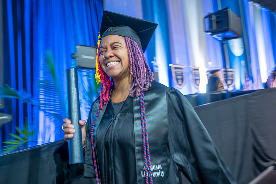 A female college graduate in cap and gown holds a diploma tube as she walks off the graduation stage during a large ceremony.