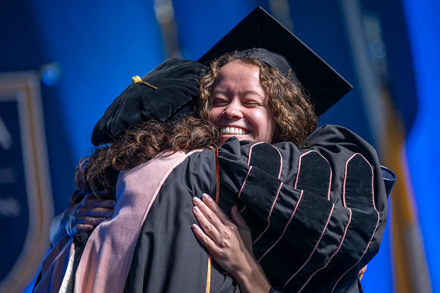 A female college graduate hugs a female professor as they celebrate graduation during a large ceremony.