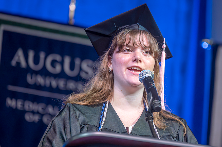 A female college graduate in cap and gown sings while standing at a podium with a microphone.