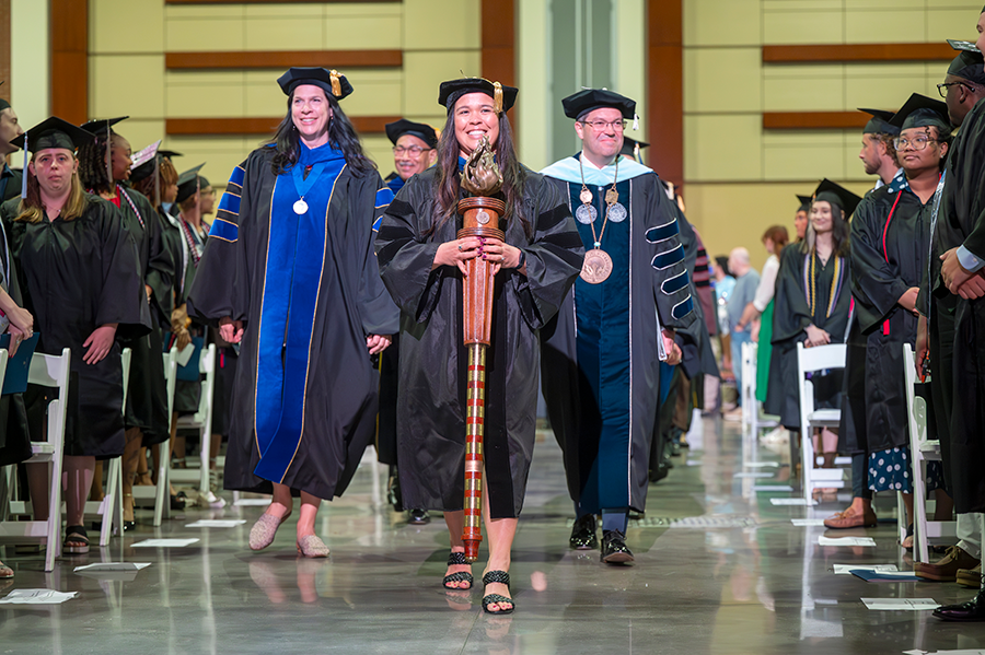 College professors wearing full graduation regalia walk down a center aisle during a graduation ceremony. The female professor in front is carrying a ceremonial mace.