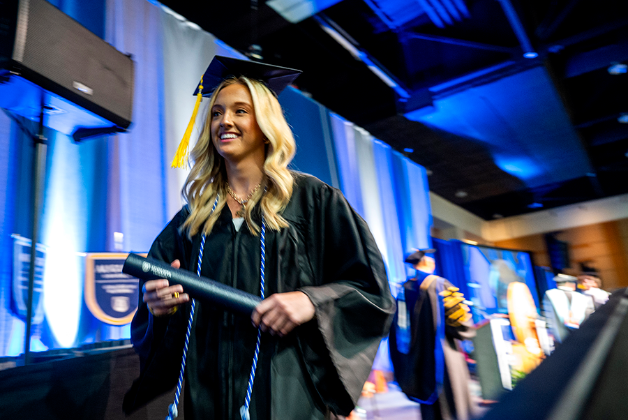 A female college graduate in cap and gown holds a diploma tube as she walks off the graduation stage during a large ceremony.