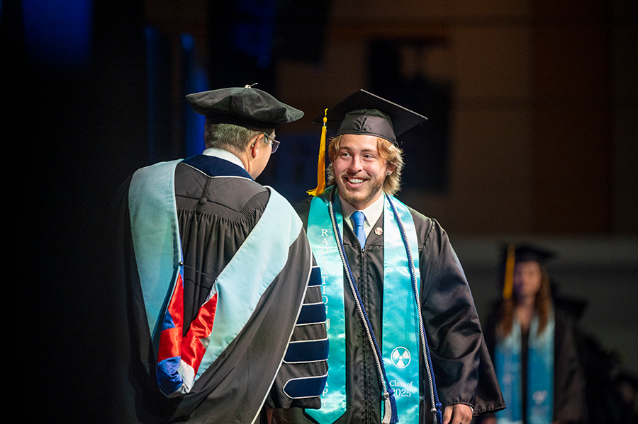 A male college graduate in cap and gown shakes the hand of a male university president during graduation.