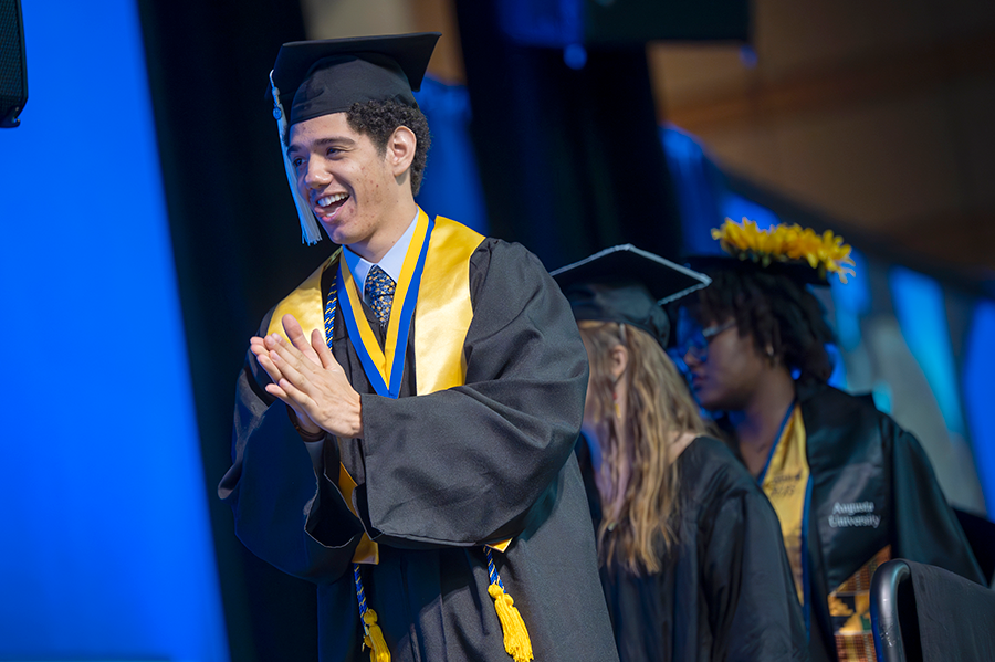 A male college graduate wearing a graduation cap and gown claps as he walks across the stage during a large graduation ceremony.