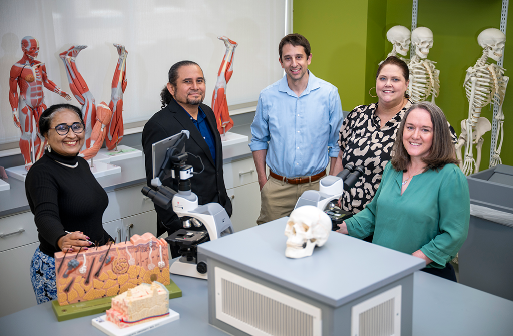 Five college professors pose in an anatomy and physiology classroom. Models of human body parts surround them.