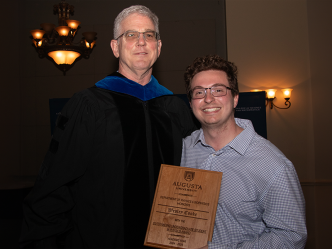 An older man wearing academic regalia stands for a photo with a student. The student holds a plaque that reads "Outstanding Undergraduate Student in Physics Award."