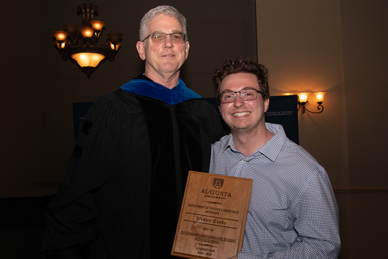 An older man wearing academic regalia stands for a photo with a student. The student holds a plaque that reads "Outstanding Undergraduate Student in Physics Award."
