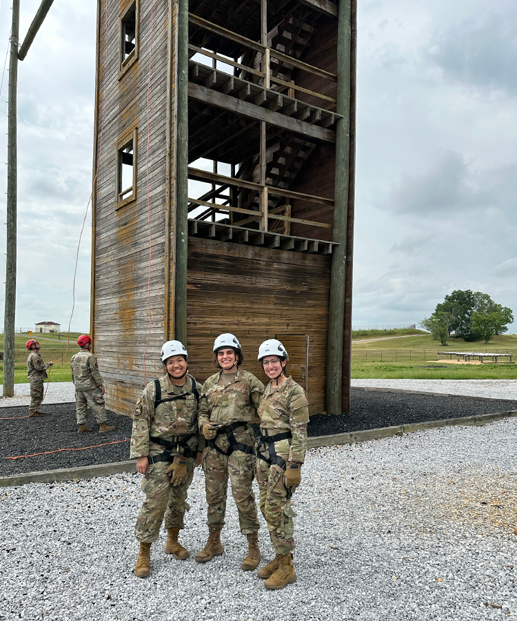Three soldiers, wearing climbing harnesses and safety hard hats, stand in front of a training climbing tower.