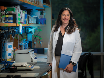 A female scientist, wearing a lab coat and holding a folder of important papers, stands in her lab.