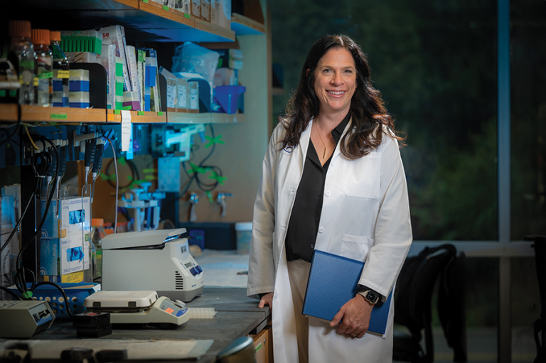 A female scientist, wearing a lab coat and holding a folder of important papers, stands in her lab.