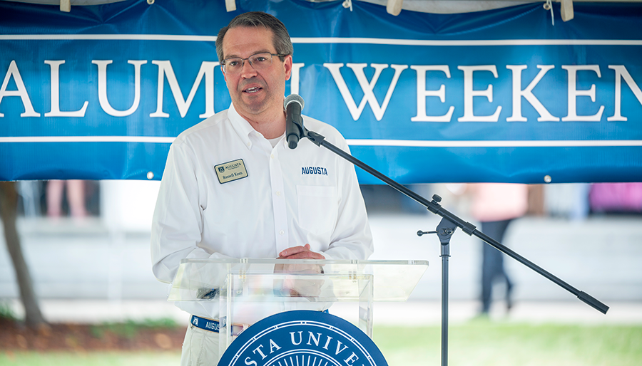 A man stands at a podium with a microphone and speaks to a large group of people during an outdoor event.