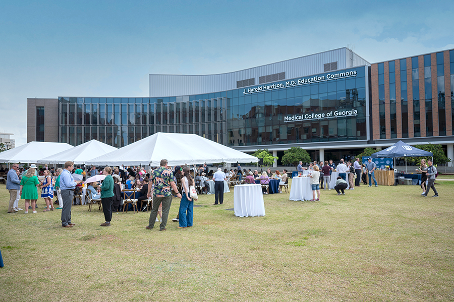 A large group of people stand around tables and chairs under and near large tents during a large event on a college campus.