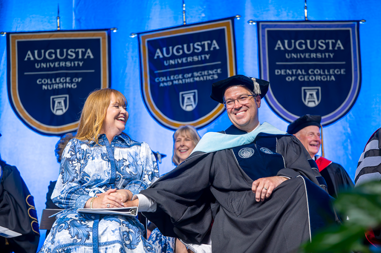 A man holds his wife's hand during a ceremony celebrating him. A large group of people clap as he is introduced.