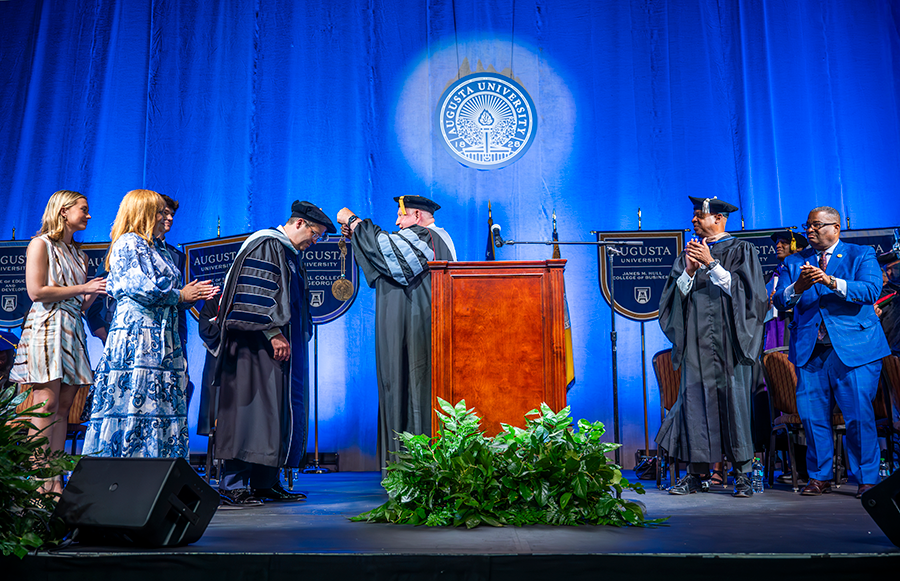 A man wearing full college graduation regalia speaks with another man, also wearing full regalia, while the second man's wife, daughter, and son, look on. They are all standing on a stage in front of a large group of people.
