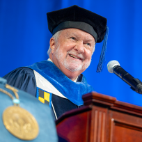 A man wearing full graduation regalia stands and speaks at a podium with a microphone during a large ceremony.