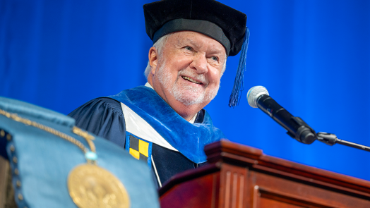 A man wearing full graduation regalia stands and speaks at a podium with a microphone during a large ceremony.