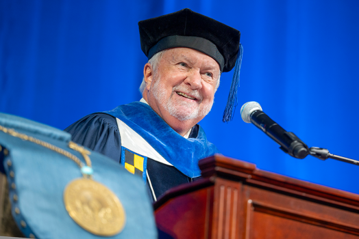 A man wearing full graduation regalia stands and speaks at a podium with a microphone during a large ceremony.