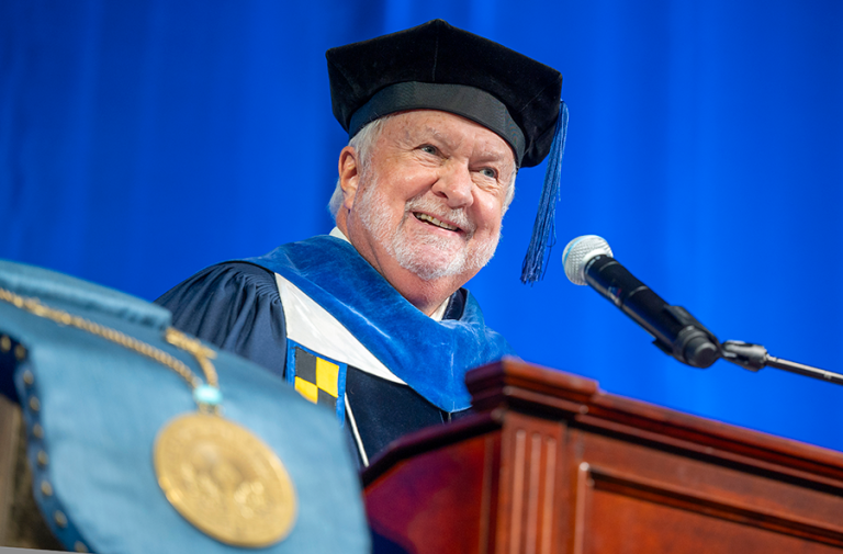 A man wearing full graduation regalia stands and speaks at a podium with a microphone during a large ceremony.
