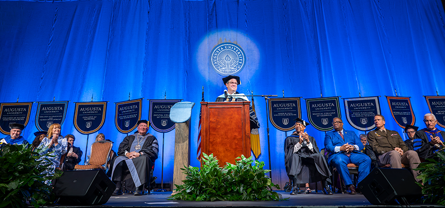 A man wearing full graduation regalia stands and speaks at a podium with a microphone during a large ceremony.