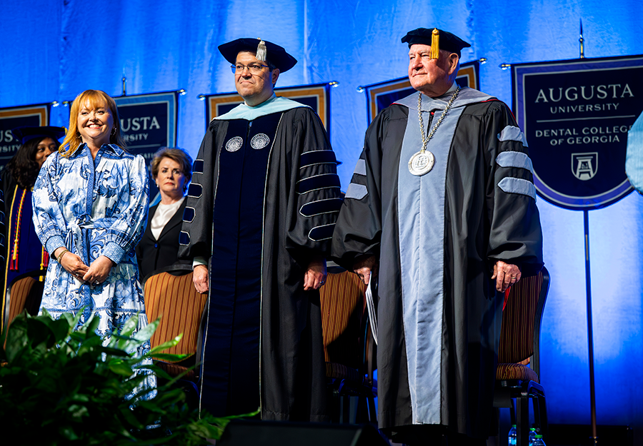 Two men wearing full college graduation regalia stand next two a woman on a stage during a large event.