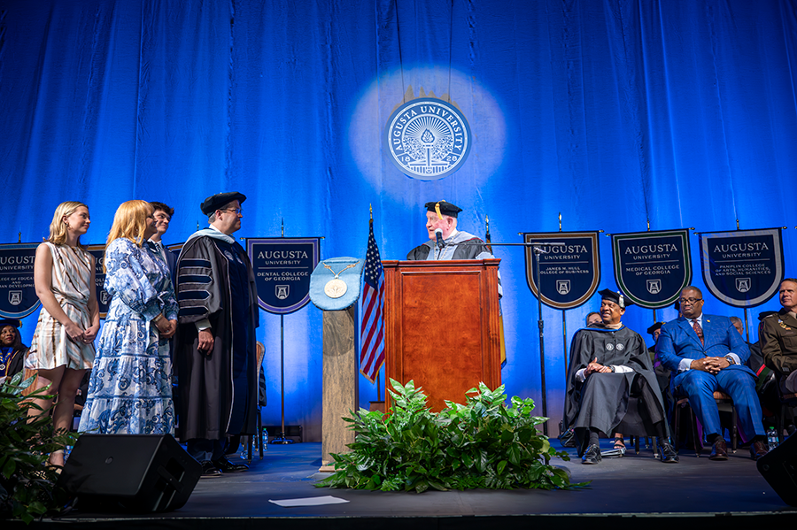 A man wearing full college graduation regalia speaks with another man, also wearing full regalia, while the second man's wife, daughter, and son, look on. They are all standing on a stage in front of a large group of people.