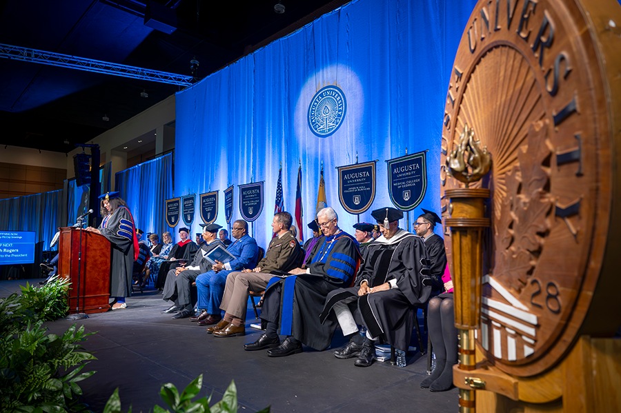 A large group of men and women, some wearing full college regalia and some wearing suits, sit on a stage in front of a large group of people during a big event.