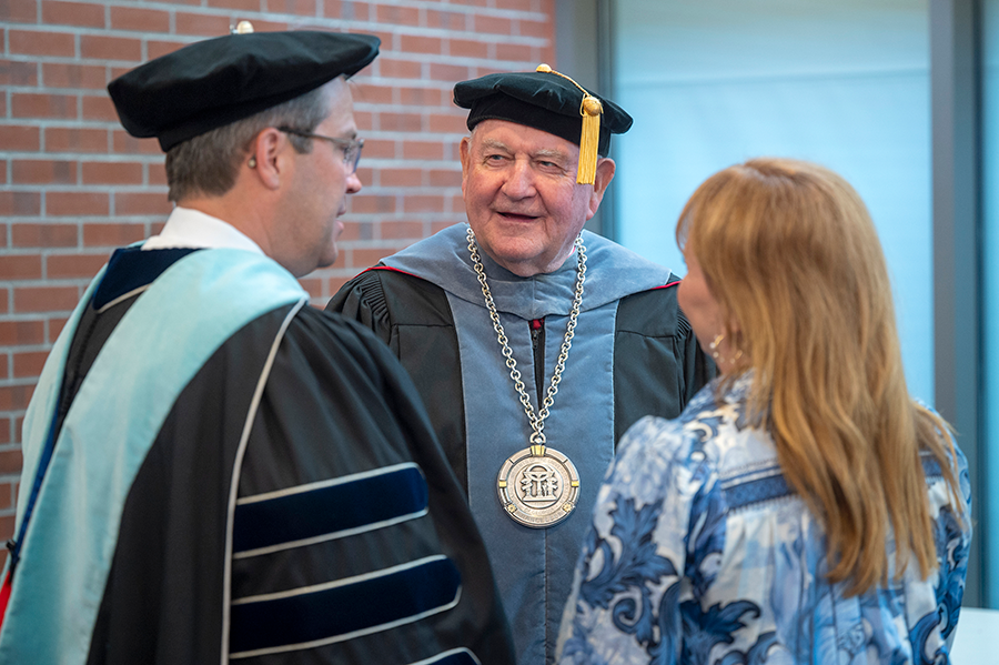 Two men wearing full graduation regalia stand and speak with a woman just before a large event.