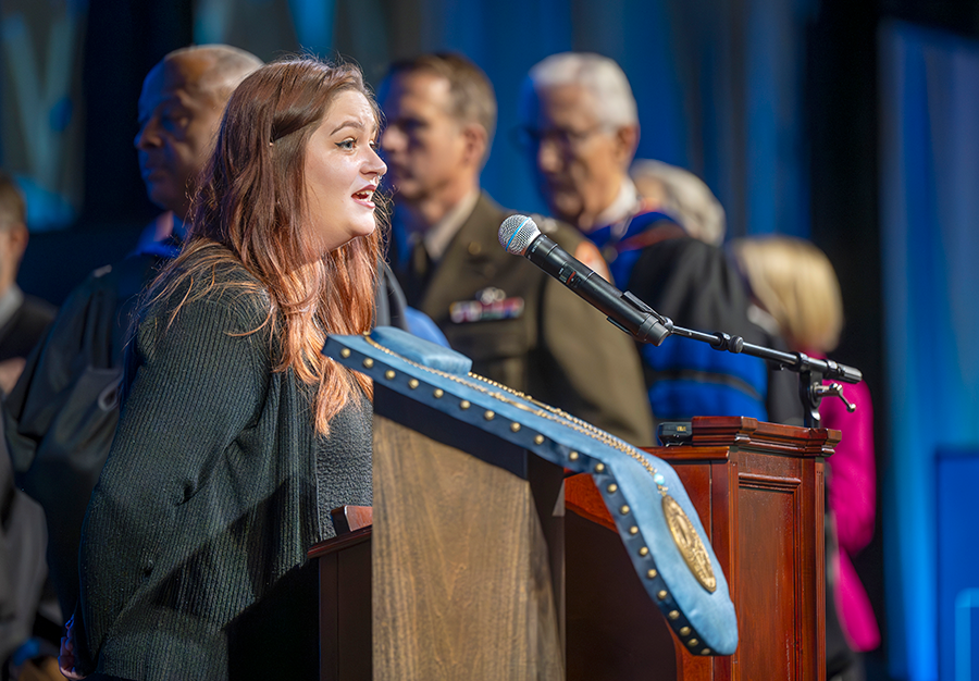 A female college student stands at a podium with a microphone and sings during a large event.