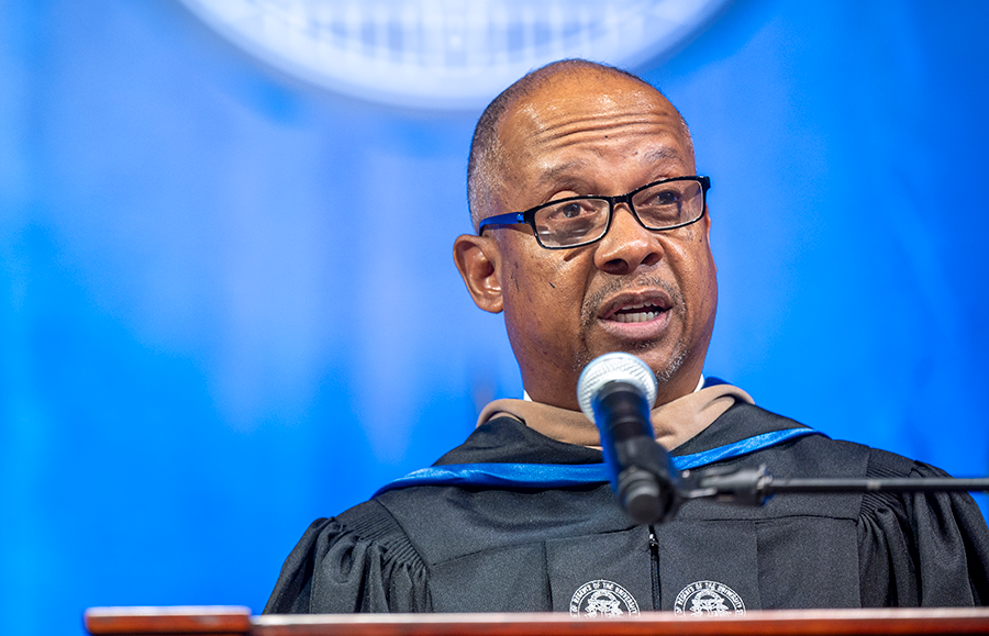A man wearing full graduation regalia stands and speaks at a podium with a microphone during a large ceremony.
