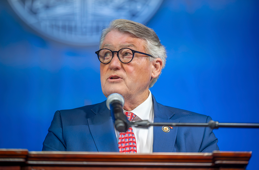 A man in a suit and tie stands at a podium with a microphone and speaks to a large group of people.
