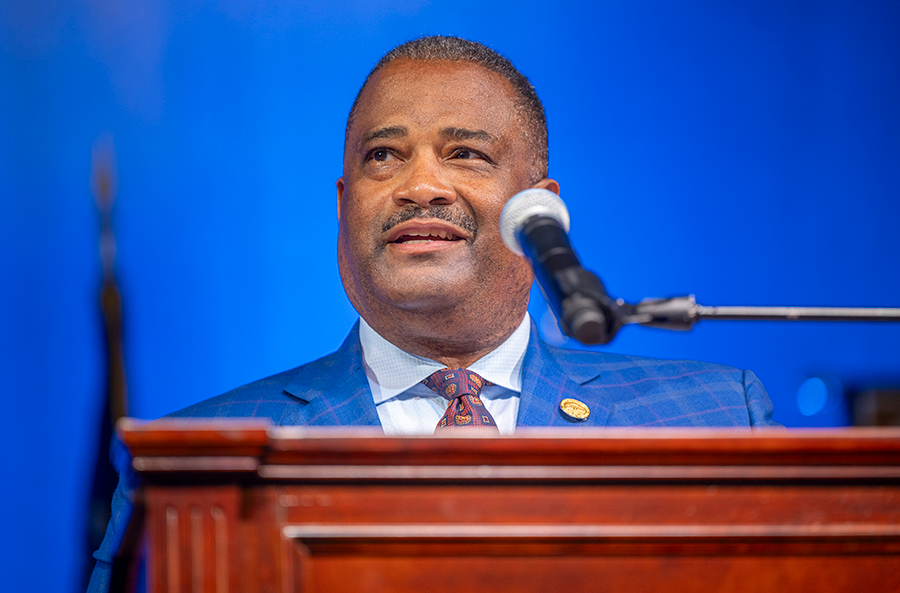 A man in a suit and tie stands at a podium with a microphone and speaks to a large group of people.