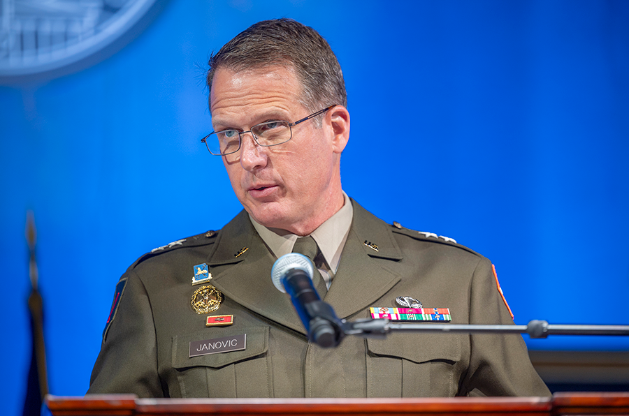 A military leader stands at a podium with a microphone and delivers a speech to a large group of people.