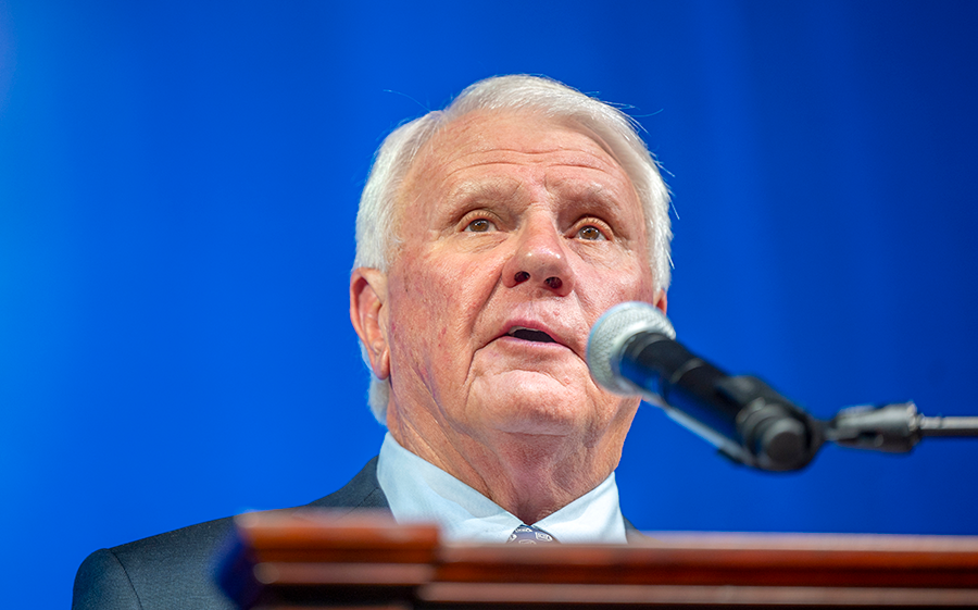 A man in a suit and tie stands at a podium with a microphone and speaks to a large group of people.