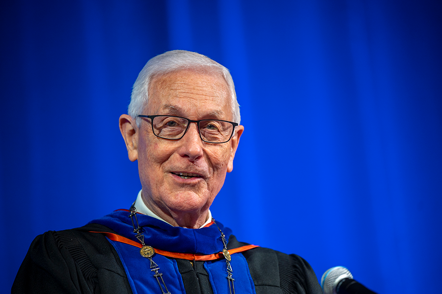 A man wearing full graduation regalia stands and speaks at a podium with a microphone during a large ceremony.