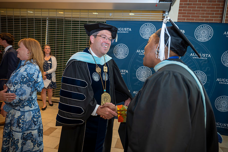 A man wearing full graduation regalia, including hood and medallion, greets a female professor, also wearing full graduation regalia, following a ceremony.