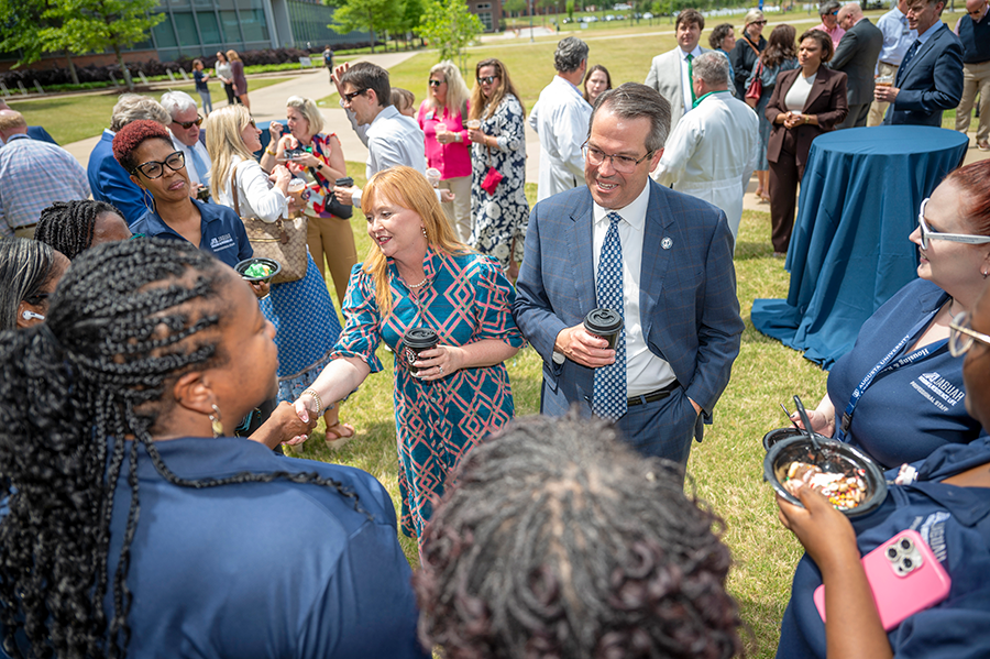 A man and a woman meet and shake hands with employees from a university. There is a large group of people around them and everyone is outside enjoying a party.