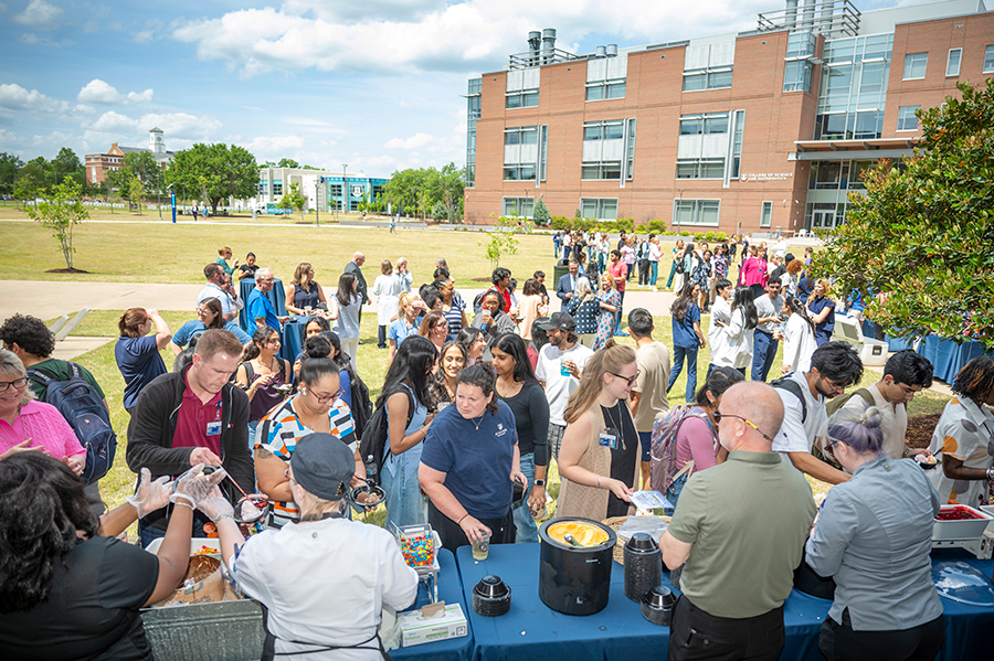 A large group of college students, faculty, and staff gather on a large lawn between several buildings on a college campus for an event.