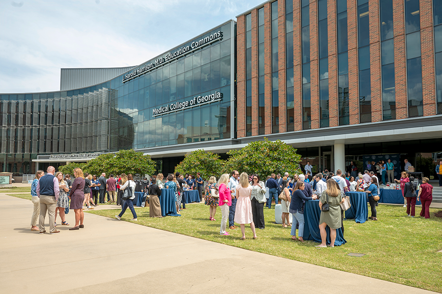 A large group of college students, faculty, and staff gather on a large lawn between several buildings on a college campus for an event.