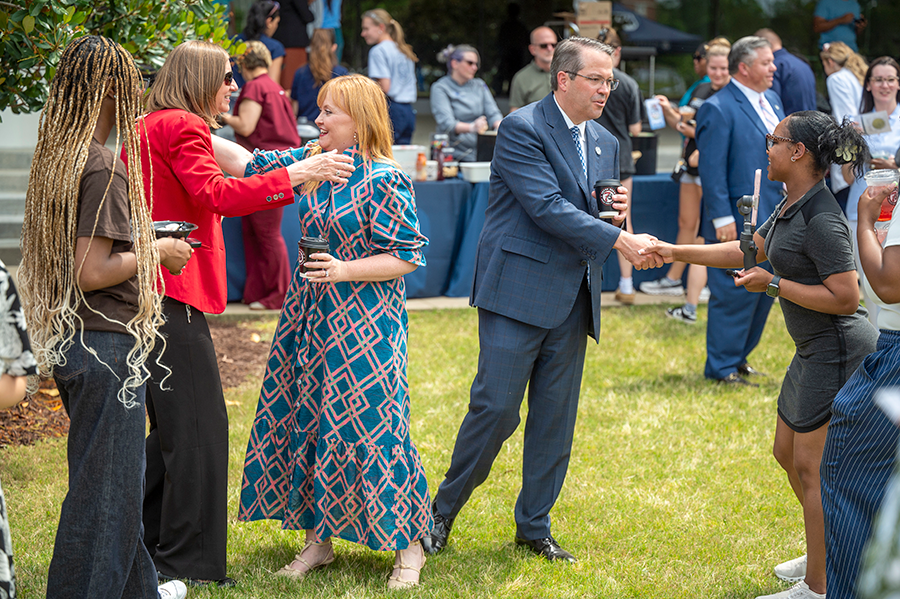 A man and a woman greet a small group of people who are part of a larger group during an outside celebration.