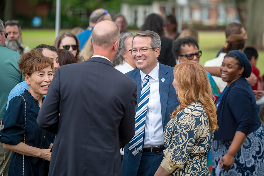A man shakes hands with another man during a large outdoor event. There is a large group of people gathered around them talking and mingling.