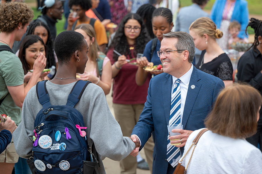 A man in a suit shakes hands with a female college student during a large outdoor event.