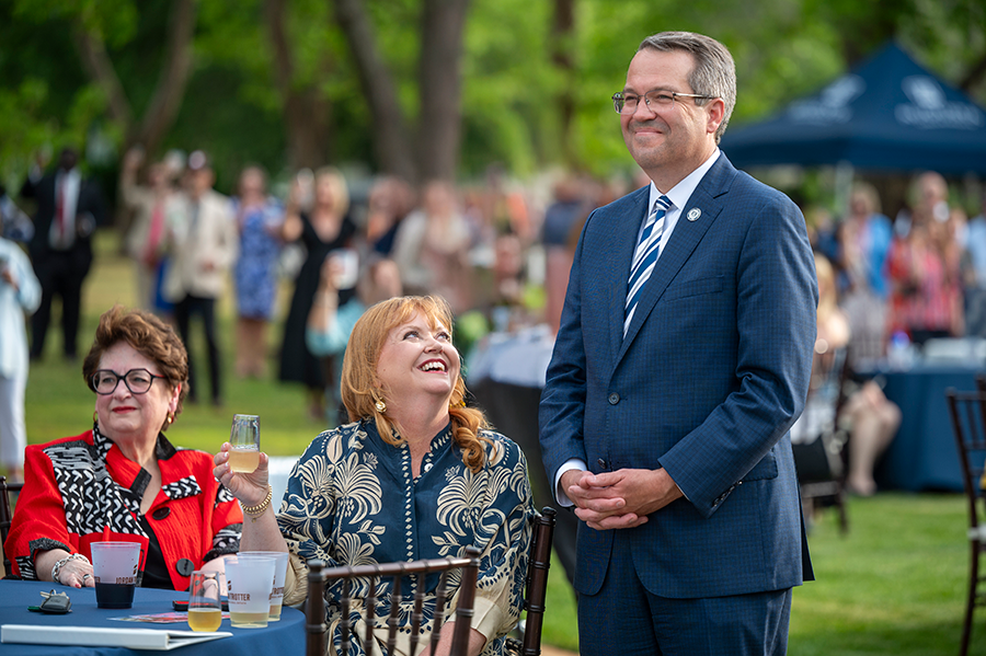 A man stands to be recognized during an outdoor event. His wife is sitting next to him and smiles as she looks in his direction. There is a large crowd gathered around tables around them.