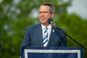 A man stands at a podium with a microphone and delivers a speech to a large group of people gathered during an outside event.