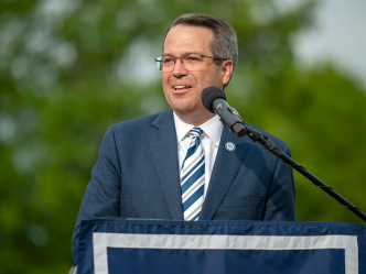 A man stands at a podium with a microphone and delivers a speech to a large group of people gathered during an outside event.
