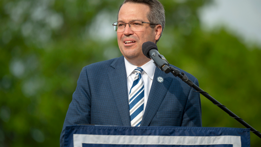 A man stands at a podium with a microphone and delivers a speech to a large group of people gathered during an outside event.