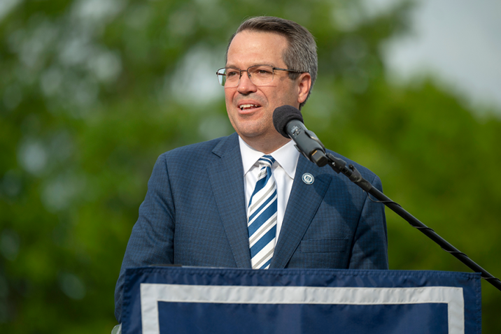 A man stands at a podium with a microphone and delivers a speech to a large group of people gathered during an outside event.