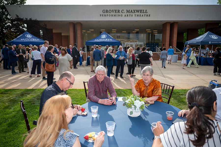 A large group of people are gathered during an event on a college campus. There are tables and chairs spread in front of a performing arts building with various people sitting around them.