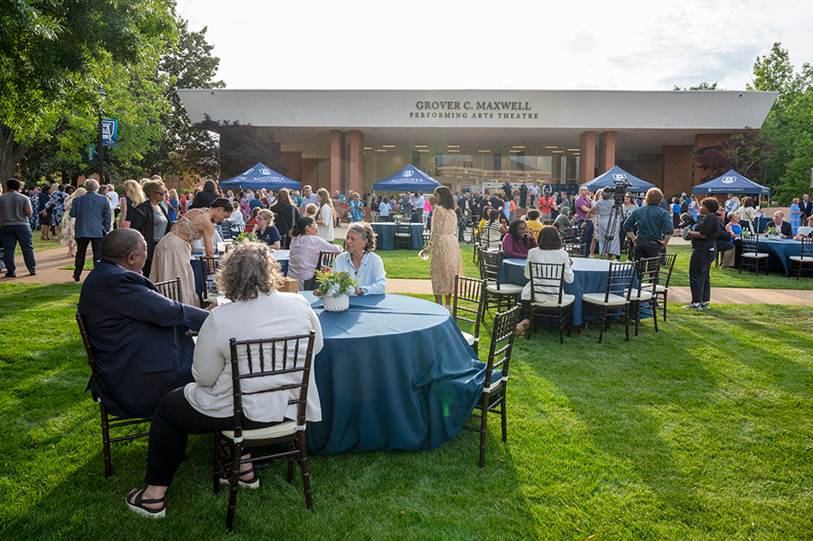 A large group of people are gathered during an event on a college campus. There are tables and chairs spread in front of a performing arts building with various people sitting around them.