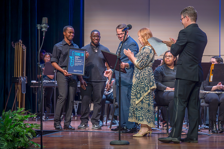 A man and his wife accept framed photos from two college students following a musical performance. There is a large band from a college seated on the stage behind them.
