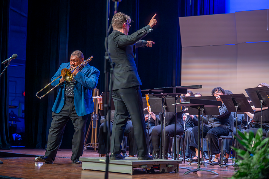 A man plays a trombone and another man directs a college wind ensemble during a performance on a stage.