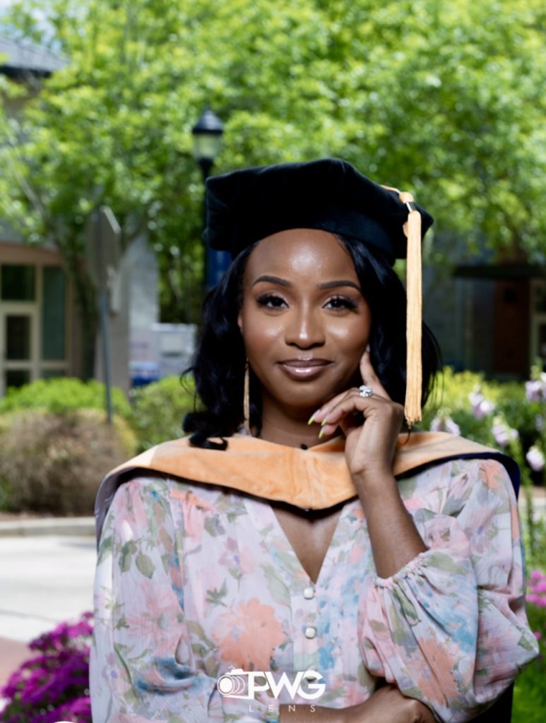 Woman in a doctoral cap and hood poses in front of a building with a large tree